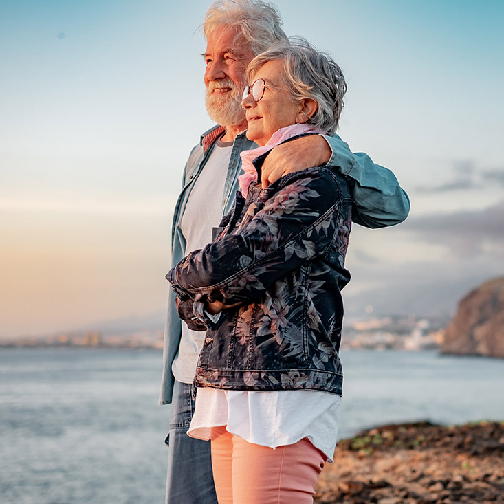 Lovely senior couple standing on the rocks at sea enjoying nature and freedom looking the horizon at sunset light.